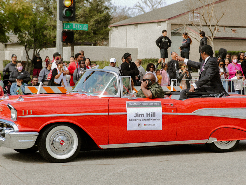 A red car with the words JIm HIll Celebrity Grand Marshall on the side as a man in the back points out to the crowd