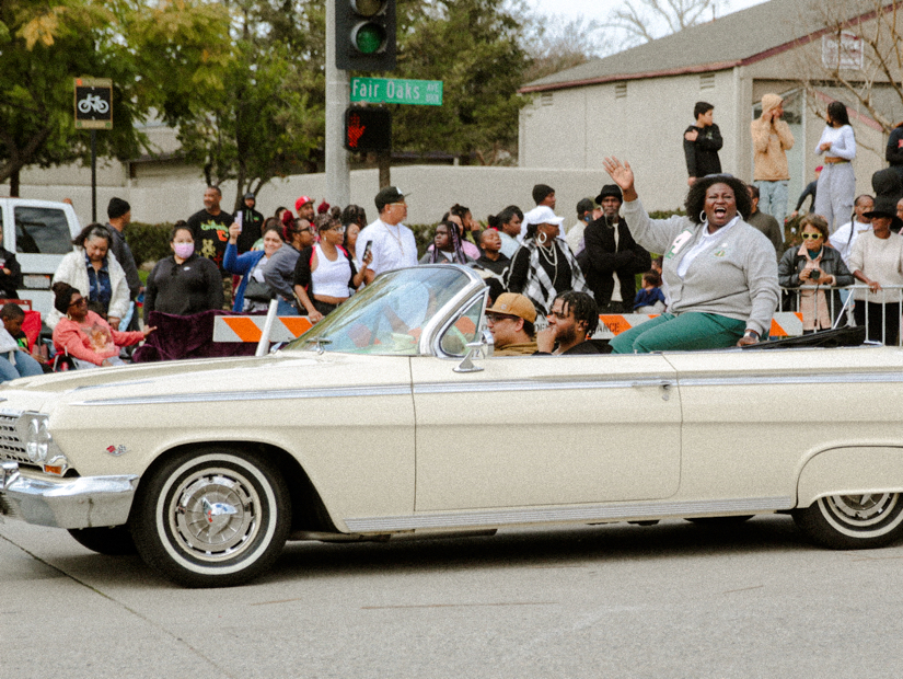 A cream white car driving down teh street of the Black History Parade
